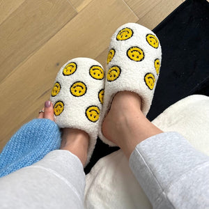 Weiße Plüsch-Hausschuhe mit gelben Smiley-Motiven, getragen an Füßen auf Holzboden | Fashion Peak